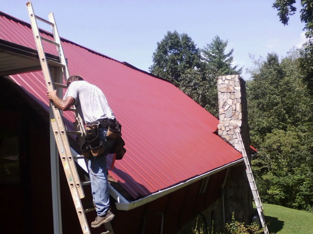 Roofer on a ladder beside a red metal roof and stone chimney — Soffit & Fascia Repair by Roofing Brothers in Porterville